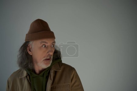 Photo for Shocked hipster style man in beanie hat and brown jacket standing with open mouth and looking away on grey background, aging population lifestyle concept, copy space - Royalty Free Image