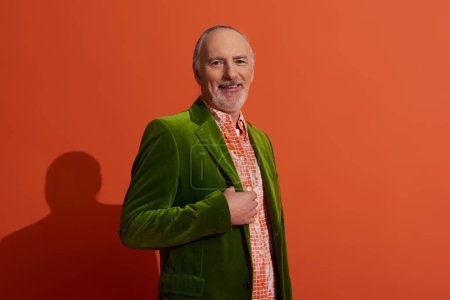 hombre de pelo gris senior, carismático y alegre, posando en camisa de moda y chaqueta de terciopelo verde sobre fondo rojo anaranjado, mirando a la cámara, sonriente, concepto de envejecimiento positivo y de moda