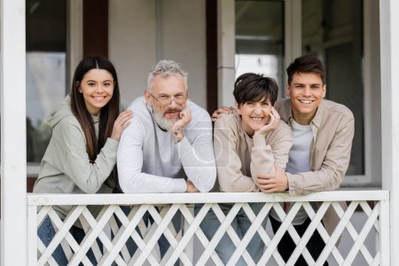 happy parents day, middle aged parents smiling next to teenage daughter and young adult son on porch of summer house, family reunion, bonding, moments to remember, modern parenting 
