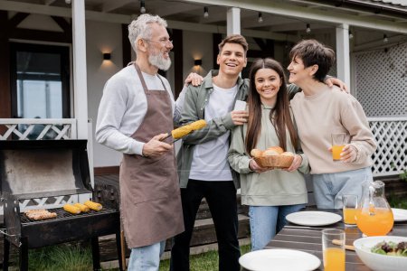 family photo, parents day, happy middle aged parents having bbq party with teenage daughter and young adult son, father holding tongs with grilled corn, summer house, suburban life 