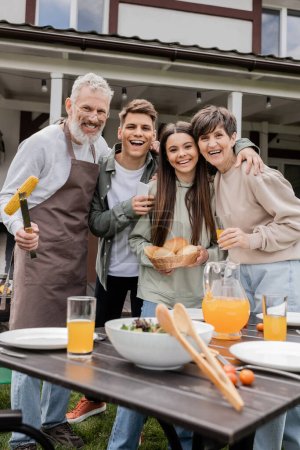 Photo for Parents day, family photo, happy middle aged parents having bbq party with young adult son and teenage daughter, father holding tongs with grilled corn, summer house, suburban life - Royalty Free Image