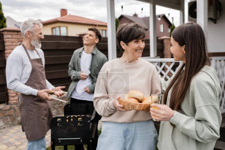 celebration of parents day, middle aged mother holding buns and talking with teenage daughter, father and son preparing food on barbecue grill, laughter, modern parenting, suburban life, summer