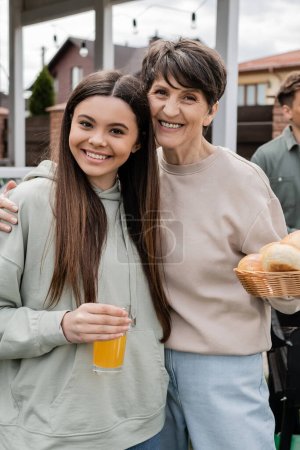 Photo for Modern parenting, celebration of parents day, happy middle aged mother holding buns and hugging cheerful teenage daughter, glass of orange juice, modern parenting, suburban life, summer - Royalty Free Image