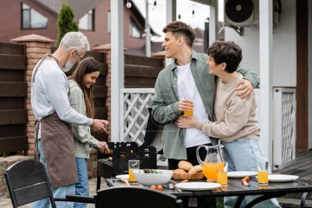Photo for Modern parenting, happy parents day, middle aged mother hugging with cheerful young adult son holding glass of orange juice, father and teenage daughter preparing food on bbq grill, summer, backyard - Royalty Free Image