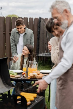 Photo for Happy teenage girl showing something on smartphone to young adult brother, digital age, father preparing food on bbq grill, barbecue party, parents day cerebration, backyard - Royalty Free Image