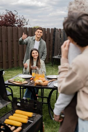 parents day celebration, happy siblings looking at father and mother on blurred background, inviting to table, gesturing, family bbq, grill party, preparing food on grill bbq, modern parenting 