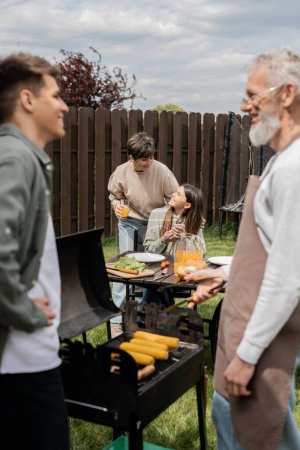 adolescent gai tenant smartphone et regardant l'âge numérique de la mère, père et fils préparant la nourriture sur barbecue grill, barbecue partie, fête des parents cérébration, arrière-cour, flou, candide 