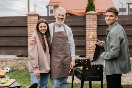 happy parents day, middle aged father hugging teenage daughter, young adult son holding tongs and grilling corn, bbq party, family photo, looking at camera, backyard of summer house 