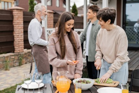 parents day, modern parenting, middle aged mother cutting lettuce near teenage daughter holding cherry tomatoes, making salad, father and son on blurred background, family bbq party