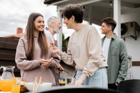 Photo for Happy middle aged mother holding cherry tomato and talking with teenage daughter, family grill party, father and son on blurred background, happy parents day concept, summer - Royalty Free Image