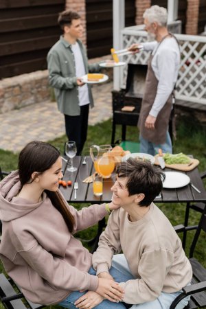 joyful teenage girl talking to middle aged mother during family grill party, summer food, father and son preparing food on bbq grill, celebration of parents day concept, backyard of house 