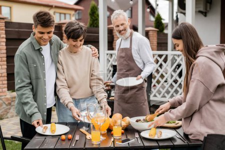 Photo for Family bbq party, celebration of parents day, happy young adult son hugging middle aged mother, bearded father in apron looking at teenage daughter, backyard of family house - Royalty Free Image