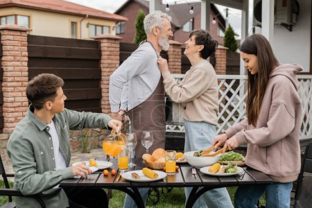 Photo for Happy middle aged couple looking at each other during family bbq party, young adult son looking at cheerful parents, love, joyful teenage girl mixing salad, happy parents day, backyard, summer - Royalty Free Image