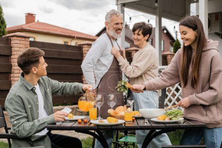 Photo for Happy middle aged couple on backyard, joyful teenage girl serving salad near adult brother pouring orange juice, celebrating parents day, love, family grill party, summer - Royalty Free Image