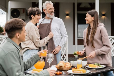 celebrating parents day, happy middle aged couple looking at joyful teenage girl serving salad near adult brother with glass of orange juice, love, family grill party, summer 