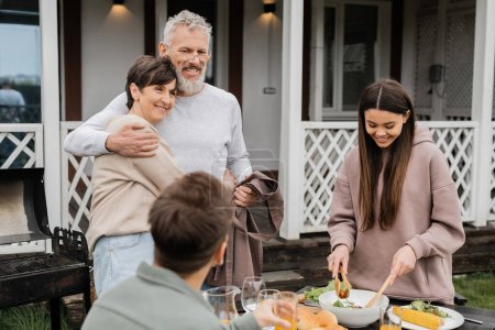 Photo for Celebrating parents day, cheerful middle aged couple hugging near joyful teenage daughter serving salad next to adult brother, love, family grill party, summer, happy marriage, summer house backyard - Royalty Free Image