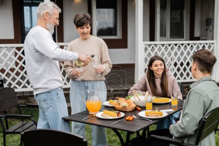 Photo for Family bbq party, middle aged man pouring wine into glass of wife near teenage daughter and adult son, grilled food, celebrating parents day on backyard of summer house, june - Royalty Free Image