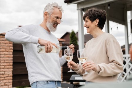 happy marriage, cheerful and bearded middle aged man in glasses pouring white wine into glass of joyful wife, backyard of summer house, romance, casual attire, spending time together 