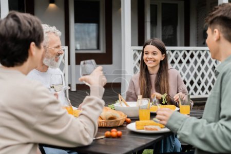 glückliches Teenagermädchen schaut fröhliche Eltern bei Familienfeiern an, sitzt auf dem Hinterhof des Sommerhauses, verbringt Zeit miteinander, isst gegrilltes BBQ-Essen, glückliches Elterntagskonzept 