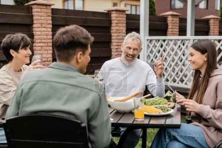 cheerful middle aged man looking at teenage daughter and gesturing during bbq party, sitting on backyard of summer house, spending time together, eating grilled bbq food, happy parents day concept 