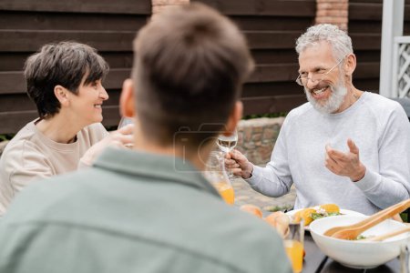cheerful middle aged man looking wife near adult son and gesturing during bbq party, blurred foreground, backyard of summer house, spending time together, eating grilled bbq food, parents day concept 