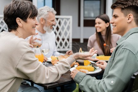 happy middle aged mother holding glass of wine and talking with adult son during bbq family party, eating grilled food, father and daughter on blurred background, backyard of summer house 
