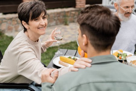 Smiling middle aged mother holding glass of wine and touching shoulder of young son near husband at background and food during bbq party and parents day celebration at backyard, family unity concept