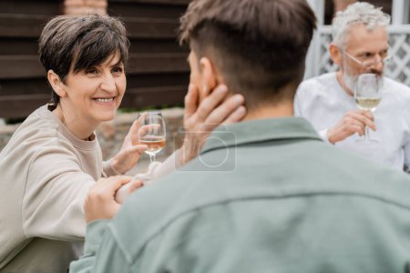 Lächelnde Mutter mittleren Alters mit einem Glas Wein und einem berührend verschwommenen erwachsenen Sohn in der Nähe des Ehemannes im Hintergrund bei der Feier zum Elterntag im Hinterhof, Familienliebe und Einheitskonzept