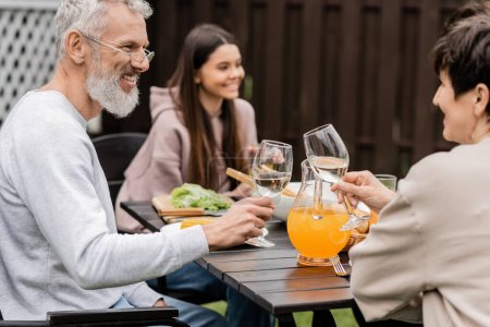 Photo for Smiling mature parents clinking glasses of wine near summer bbq food and blurred teenage daughter during parents day celebration at backyard, family love and unity concept, tradition and celebration - Royalty Free Image