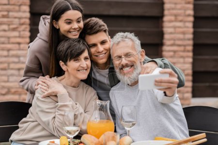 Téléchargez les photos : Sourire papa d'âge moyen prendre selfie sur smartphone avec la famille et les enfants près de savoureux repas d'été lors de la fête barbecue et fête des parents à l'arrière-cour, l'amour de la famille et concept d'unité - en image libre de droit