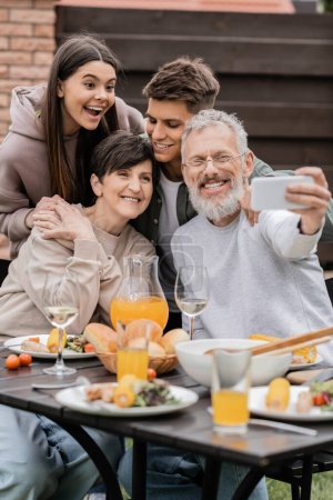 Téléchargez les photos : Père mature positif prenant selfie sur smartphone avec sa femme et ses enfants près de la nourriture d'été floue pendant la fête barbecue et la fête des parents à la cour, concept heureux de la journée des parents - en image libre de droit