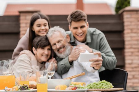 Smartphone in der Hand eines verschwommenen reifen Mannes, der ein Selfie mit Familie und Kindern beim sommerlichen Essen während der Grillparty und der Elterntagsfeier im Hinterhof im Juni macht, glückliches Konzept für den Elterntag