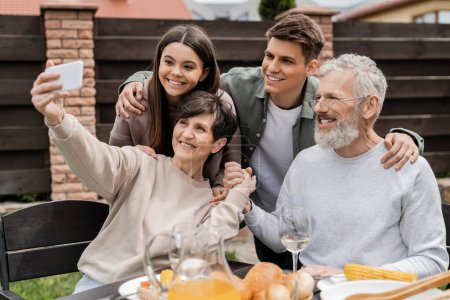 Smiling middle aged woman holding hand of husband while taking selfie on smartphone with family and kids during bbq party and parents day celebration at backyard, happy parents day concept