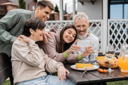Positive teenage girl using smartphone near mature parents and father during bbq party with summer food and parents day celebration at backyard in june, happy parents day concept, special occasion