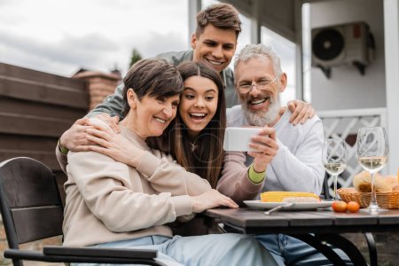 Photo for Cheerful teenage girl holding smartphone near middle aged parents and young brother during bbq summer party and parents day celebration at backyard in june, happy parents day concept - Royalty Free Image