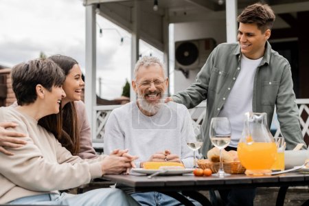 Positive Frau und Kinder sehen lachenden Mann mittleren Alters beim Sommeressen während Grillparty und Elterntagsfeier im Juni, glückliches Elterntagskonzept