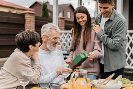 Photo for Smiling siblings giving present box to middle aged parents near summer food during barbeque party and parents day celebration at backyard in june, celebrating parenthood day concept - Royalty Free Image