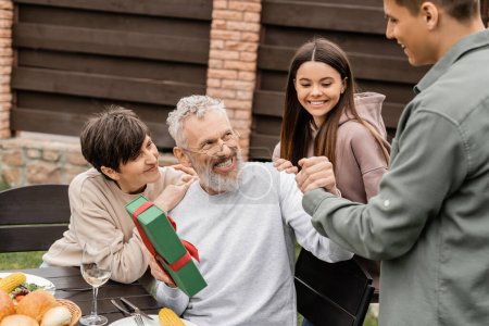 Cheerful middle aged father shaking hand of young son near wife and daughter while holding gift box during bbq party and parents day celebration at backyard, celebrating parenthood day concept