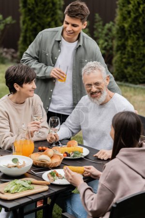 Photo for Smiling kids talking to middle aged parents near tasty summer food and parents day celebration during barbeque party at backyard in june, cherishing family bonds concept, special occasion - Royalty Free Image