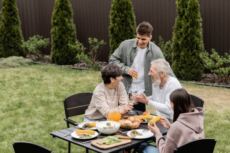 Aufgeregte und gut gelaunte Eltern mittleren Alters stoßen bei Grillparty im Hinterhof mit Weingläsern und sommerlichem Essen an, pflegen Familienbande, verbringen Zeit miteinander