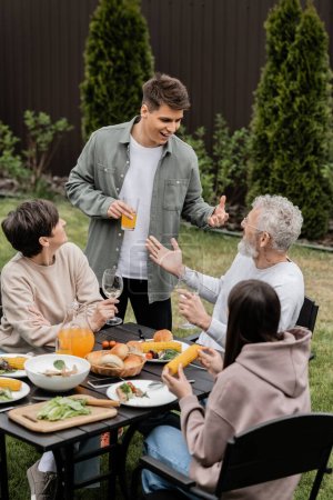 Hombre joven y sonriente sosteniendo jugo de naranja y hablando con un padre de mediana edad cerca de la familia durante la fiesta de barbacoa y la celebración del día de los padres en el patio trasero, apreciando el concepto de lazos familiares