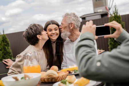 Mature parents kissing cheerful teenage daughter while son taking photo on smartphone during summer bbq party and parents day celebration at backyard, special day for parents concept