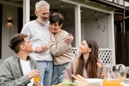 Lächelnder älterer Mann umarmt Frau und hält Wein in der Hand und spricht mit Kindern in der Nähe von sommerlichem Essen während der Grillparty und des Elterntags im Hinterhof, besonderer Tag für Eltern