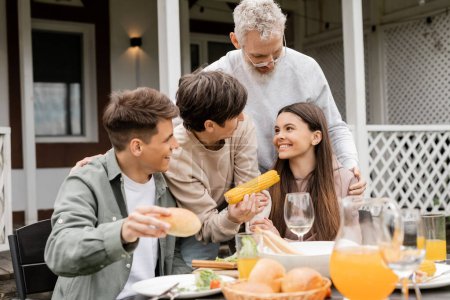 Smiling middle aged parents hugging teen daughter near son and tasty food during bbq party and parents day celebration at backyard in june, quality time with parents concept 