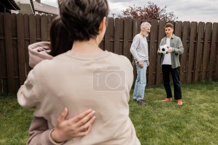 Middle aged man standing near young son with football near blurred wife and daughter during parents day celebration at backyard, quality time with parents concept, tradition and celebration
