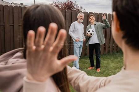 Smiling young man holding football and gesturing at sister and mom and standing near middle aged father at backyard during parents day celebration in june, quality time with parents concept 