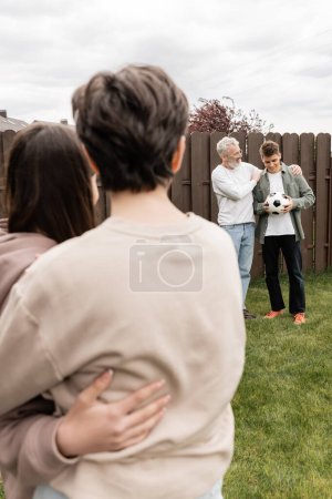 Padre de mediana edad abrazando hijo pequeño con el fútbol cerca de la familia borrosa en el patio trasero durante la celebración del día de los padres en junio, tiempo de calidad con el concepto de los padres, ocasión especial