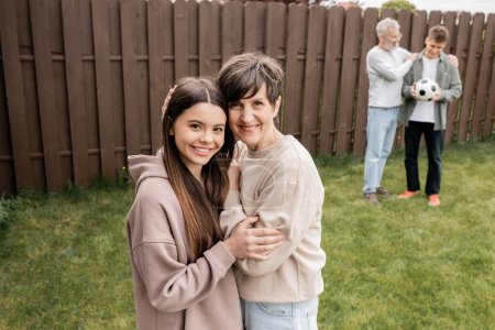 Smiling middle aged woman hugging teenage daughter and looking at camera near blurred family with football during parents day celebration at backyard, quality time with parents concept 