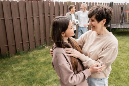 Positive teenage girl hugging middle aged mom near blurred family with football during parents day celebration at backyard in june, quality time with parents concept, tradition and celebration
