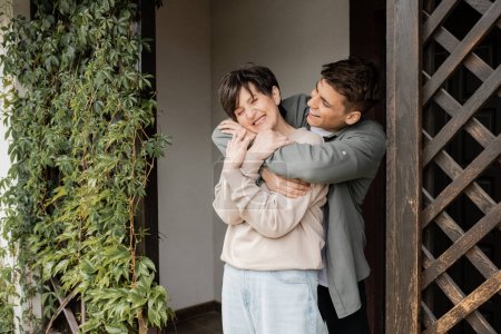 Positive young man hugging cheerful middle aged mother and standing on porch of house during parents day celebration in june, family traditions and celebrations concept, special occasion